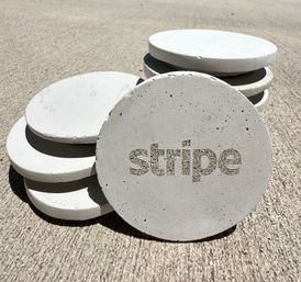 Sunlit stack of round white concrete coasters on textured outdoor pavement, front disc featuring a stenciled word cutout