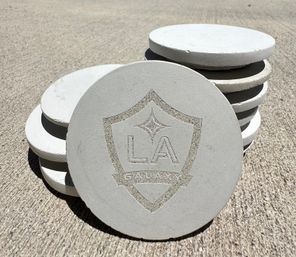 Sunlit stack of round concrete coasters with an engraved shield emblem and lettering, arranged on rough outdoor concrete pavement