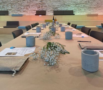 Long communal table set for a workshop in a modern industrial event space with exposed white brick wall, paper-covered tabletop lined with baby’s-breath floral runners, concrete votive holders, pens, papers and fabric pouches