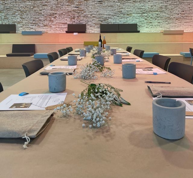 Long communal table set for a workshop in a modern industrial event space with exposed white brick wall, paper-covered tabletop lined with baby’s-breath floral runners, concrete votive holders, pens, papers and fabric pouches