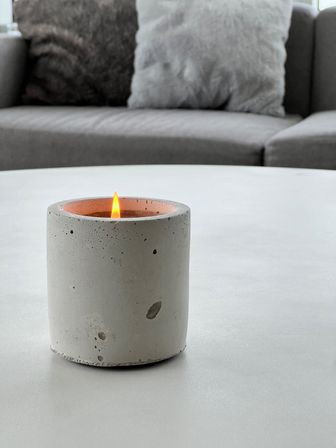 Lit candle in a cylindrical concrete holder on a white coffee table with a grey sofa and fuzzy pillows in a cozy minimalist living room