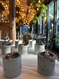 Concrete candle holders with long wicks arranged on a white tablecloth in a cozy bistro patio with hanging string lights and wicker chairs by glass windows