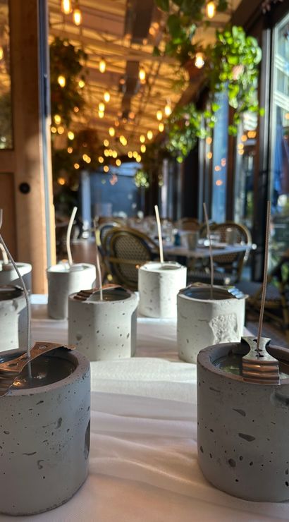 Concrete candle holders with long wicks arranged on a white tablecloth in a cozy bistro patio with hanging string lights and wicker chairs by glass windows
