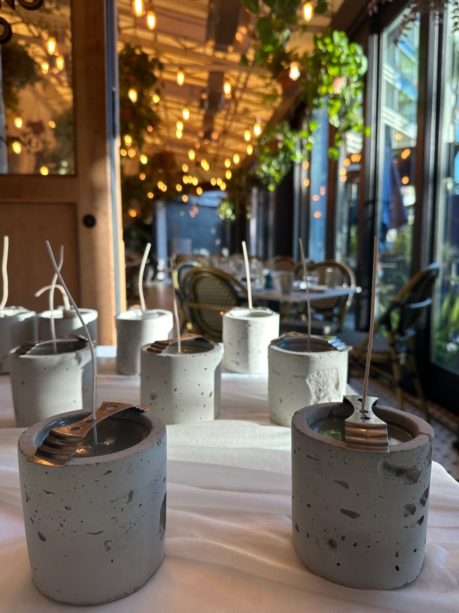 Concrete candle holders with long wicks arranged on a white tablecloth in a cozy bistro patio with hanging string lights and wicker chairs by glass windows