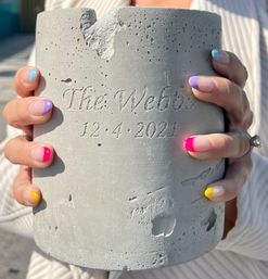 Hands with colorful French-tip manicure holding a textured concrete cylinder engraved with cursive lettering and the carved date 12·4·2021.