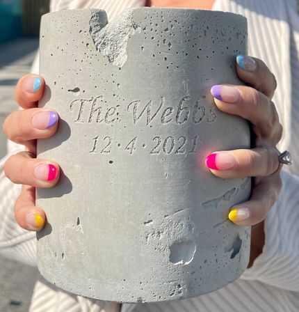 Hands with colorful French-tip manicure holding a textured concrete cylinder engraved with cursive lettering and the carved date 12·4·2021.