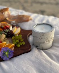 Concrete candle in a textured holder on a white blanket beside a wooden charcuterie board with grapes, orange slices, cheese and bread at a beach picnic.