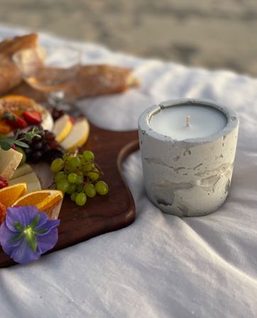 Concrete candle in a textured holder on a white blanket beside a wooden charcuterie board with grapes, orange slices, cheese and bread at a beach picnic.