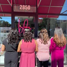 Four women stand with their backs to the camera outside a pink-awning salon storefront, wearing black and pink outfits and showing glossy curled hairstyles; one sports a small white floral hair accessory.