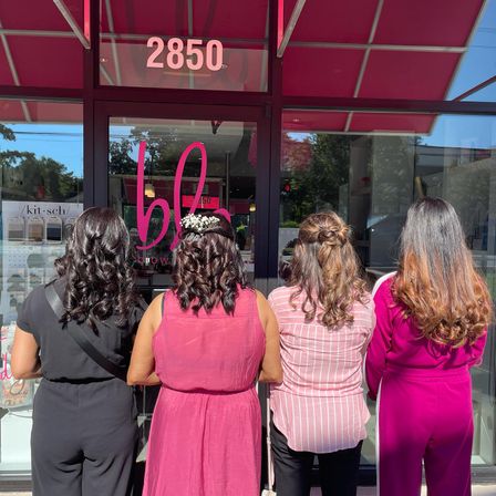 Four women stand with their backs to the camera outside a pink-awning salon storefront, wearing black and pink outfits and showing glossy curled hairstyles; one sports a small white floral hair accessory.
