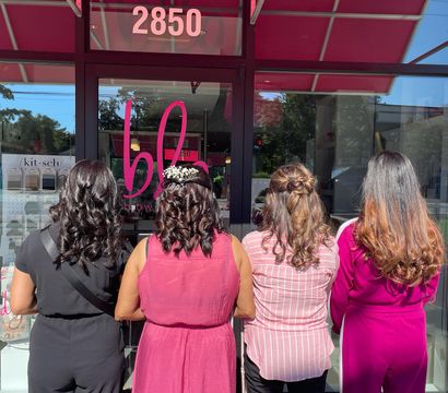 Four women stand with their backs to the camera outside a pink-awning salon storefront, wearing black and pink outfits and showing glossy curled hairstyles; one sports a small white floral hair accessory.