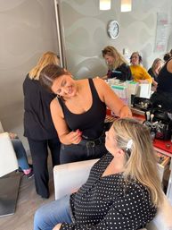 Smiling makeup artist applies eyeliner to a seated client in a modern beauty salon, makeup brushes, palettes and a mirrored station with other stylists visible in the background.