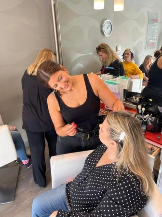 Smiling makeup artist applies eyeliner to a seated client in a modern beauty salon, makeup brushes, palettes and a mirrored station with other stylists visible in the background.
