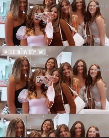 Six friends taking a mirror selfie inside a bright hair blowout salon, smiling in summer outfits with handbags and a dangling face mask — fun salon selfie scene.