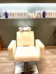 Cream salon wash chair with attached shampoo bowl and row of haircare product bottles on a light green mosaic tile wall