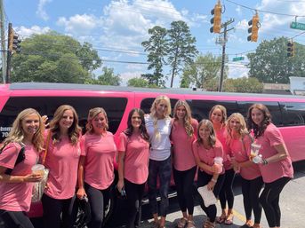 Group of women in matching pink t-shirts smiling and holding drinks in front of a pink stretch limousine on a sunny suburban street with trees and traffic lights, girls' day out.