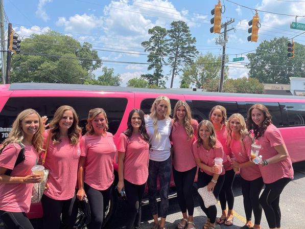 Group of women in matching pink t-shirts smiling and holding drinks in front of a pink stretch limousine on a sunny suburban street with trees and traffic lights, girls' day out.