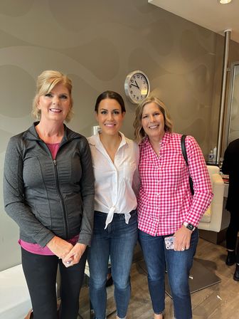 Three smiling women in casual jeans and tops pose for a group photo inside a modern salon-style interior with wavy wallpaper and a wall clock.