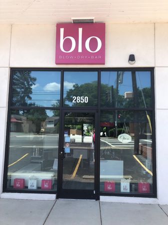 Glass-front blow-dry bar storefront with bright pink square sign, black-framed door and reflective windows showing trees and street, hanging "Open" sign and sidewalk in front.