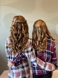 Two girls shown from behind with long golden-brown curls in half-up twisted styles, wearing coordinating plaid shirts and hugging—cozy plaid outfits and cascading curl hairstyle inspiration.