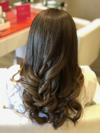 Back view of long shiny chestnut-brown hair styled in soft salon curls on a white chair