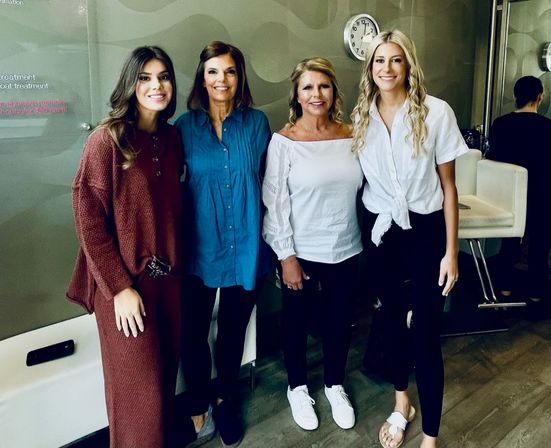 Four smiling women standing side-by-side inside a modern salon or spa, casual outfits, styling chairs and a wall clock visible in the background.