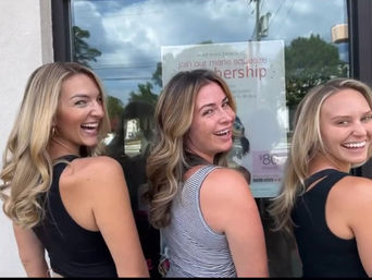 Three laughing women with beachy blonde and highlighted waves in sleeveless tops posing over their shoulders outside a hair salon window with a membership poster