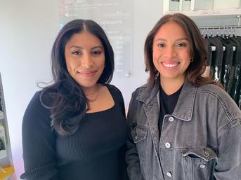 Two smiling women with styled hair posing inside a modern hair salon, one in a black top and one in a gray denim jacket near hanging garment bags and a price board.