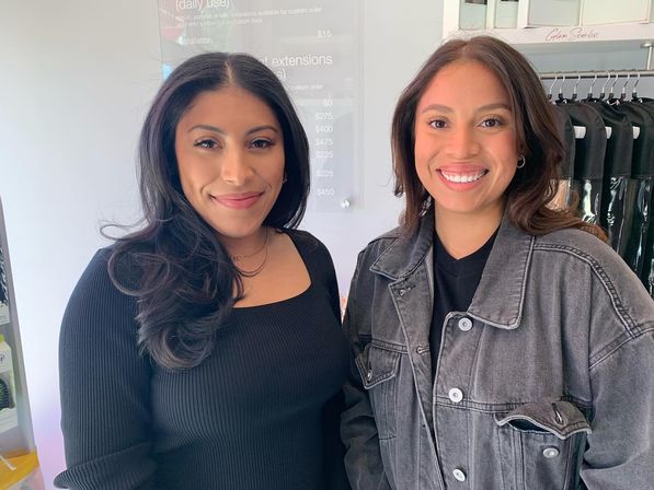 Two smiling women with styled hair posing inside a modern hair salon, one in a black top and one in a gray denim jacket near hanging garment bags and a price board.