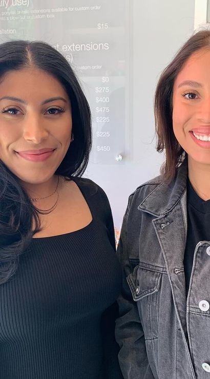Two smiling women with styled hair posing inside a modern hair salon, one in a black top and one in a gray denim jacket near hanging garment bags and a price board.