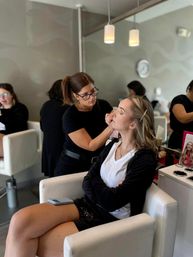 Makeup artist applying eye makeup to a seated client in a modern salon with white chairs, mirrored wall and pendant lights — relaxed getting-ready scene.