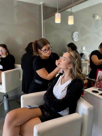 Makeup artist applying eye makeup to a seated client in a modern salon with white chairs, mirrored wall and pendant lights — relaxed getting-ready scene.