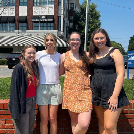 Four smiling young women in casual summer outfits standing arm‑in‑arm on a sunny downtown sidewalk in front of a brick ledge, striped awning and multi‑story storefront.