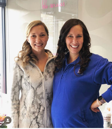 Two smiling women with styled loose curls posing inside a hair salon in front of a visible hair menu sign, casual outfits and friendly vibe.