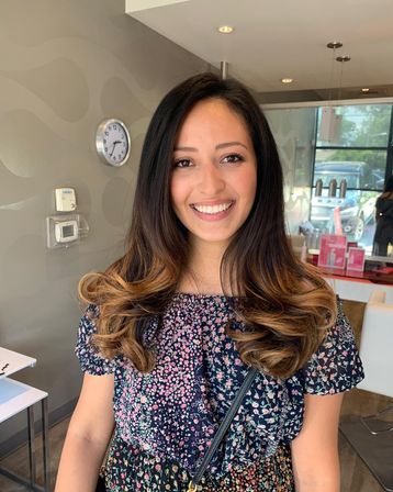 Smiling woman with long dark-to-caramel ombré hair styled in soft curls, wearing a floral dress inside a bright hair salon with styling products and a wall clock in the background.