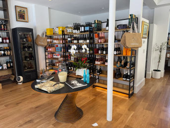 Cozy boutique wine shop interior with wooden floors, black metal shelving filled with wine and sparkling bottles, a refrigerated display, hanging canvas tote bags, and a round central table topped with an orchid and gift items.