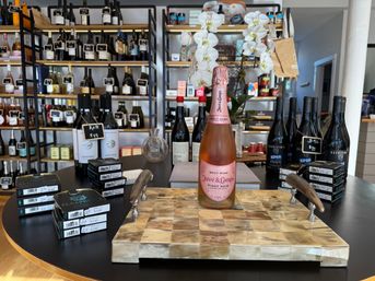 Sparkling Brut Rosé Pinot Noir bottle showcased on a checkered wooden board in a boutique wine shop, surrounded by stacked wine boxes, shelving of bottles and white orchids.