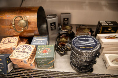 Retail shelf of gourmet canned seafood: stacked net-wrapped round tins and boxed tuna and spider-crab pâté displayed beside a vintage copper pot.