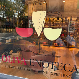 Wine shop window with playful decals of red, white and sparkling wine glasses, cozy leather seating, shelves of bottles and wooden crates inside, and street reflections on the glass.