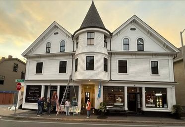 Historic white Victorian-style three-story building with a conical turret and steep gables on a downtown corner, ground-floor storefronts with ladders and people working outside at sunset.