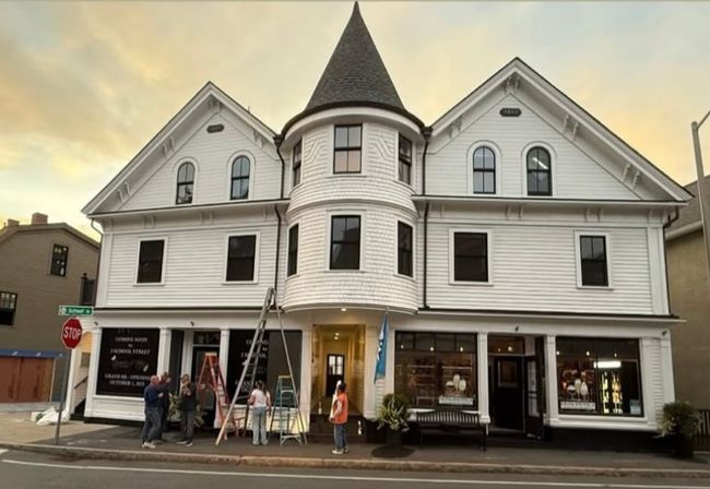 Historic white Victorian-style three-story building with a conical turret and steep gables on a downtown corner, ground-floor storefronts with ladders and people working outside at sunset.