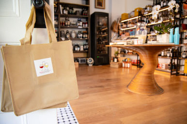 Canvas reusable tote bag with a wine-glass logo hanging on a door knob in a cozy boutique wine shop interior, wooden spiral display table and shelves stocked with wine bottles and gourmet products