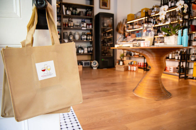 Canvas reusable tote bag with a wine-glass logo hanging on a door knob in a cozy boutique wine shop interior, wooden spiral display table and shelves stocked with wine bottles and gourmet products