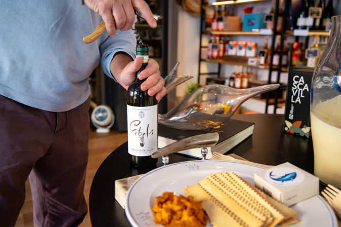 Hands twisting a corkscrew into a red wine bottle on an artisan shop counter, with a glass decanter, plate of crackers and spread, and shelves of gourmet products in the background, ready for tasting.