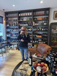 Person tasting red wine inside a cozy wine shop tasting room with floor-to-ceiling wine shelves, leather armchair, wooden floors, and a table of bottles, glasses and apples.