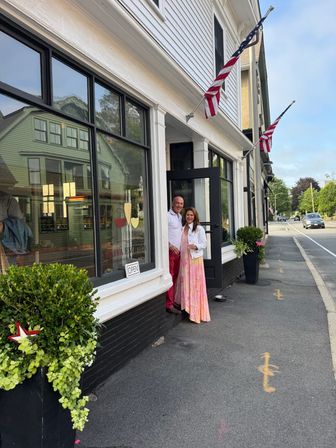 Cheerful couple standing in the doorway of a small-town storefront with large display windows, potted plants, American flags, and an 'OPEN' sign.