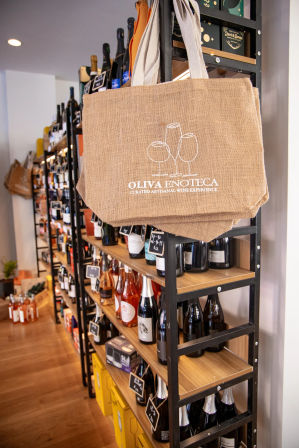 Jute tote bag with white wine-glass graphic hanging on metal-and-wood shelving stocked with assorted wine bottles in a cozy boutique wine shop interior.