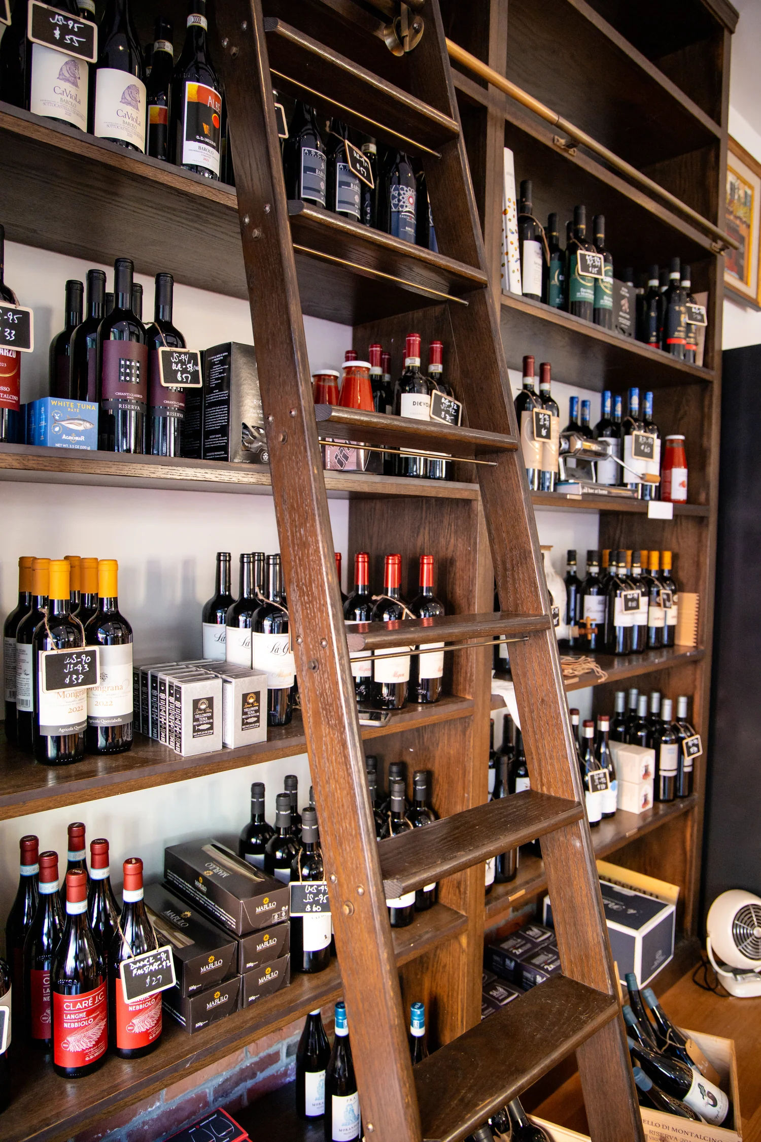 Cozy boutique wine shop interior with a sliding wooden ladder leaning against dark wood shelves stocked with assorted red and white wine bottles, boxed wines, and small chalk price tags.