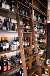 Cozy boutique wine shop interior with a sliding wooden ladder leaning against dark wood shelves stocked with assorted red and white wine bottles, boxed wines, and small chalk price tags.
