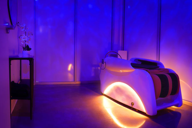 Futuristic white relaxation pod in a dim spa room with blue-purple ambient lighting, warm orange underglow and an orchid on a side table